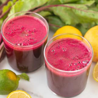 two glasses of homemade beet juice surrounded by lemons and beet greens.