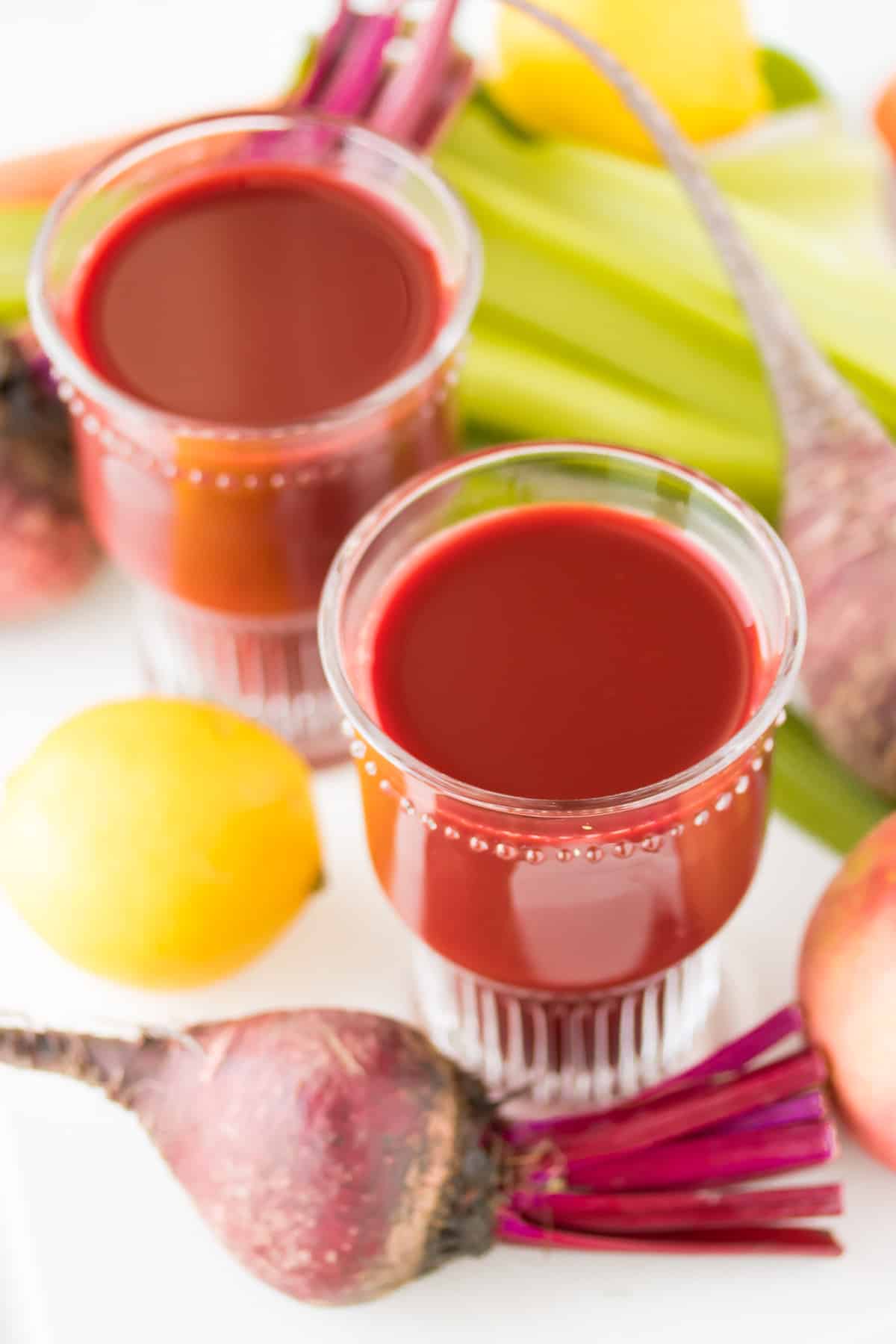 two glasses of beet juice surrounded by fresh vegetables and lemons.