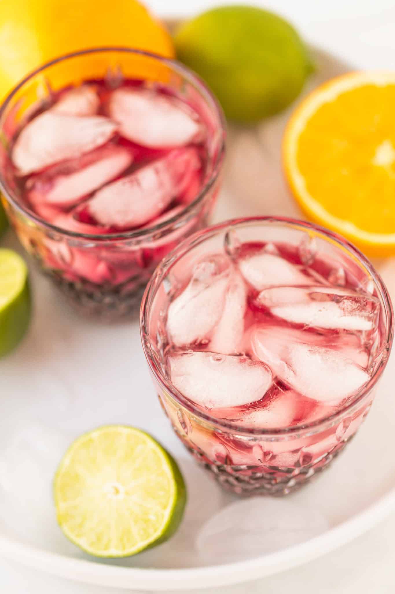 two tart cherry mocktail glasses with ice on a platter with oranges and limes
