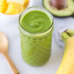 detox smoothie in a glass surrounded by a spoon, bananas, avocado, and pineapple