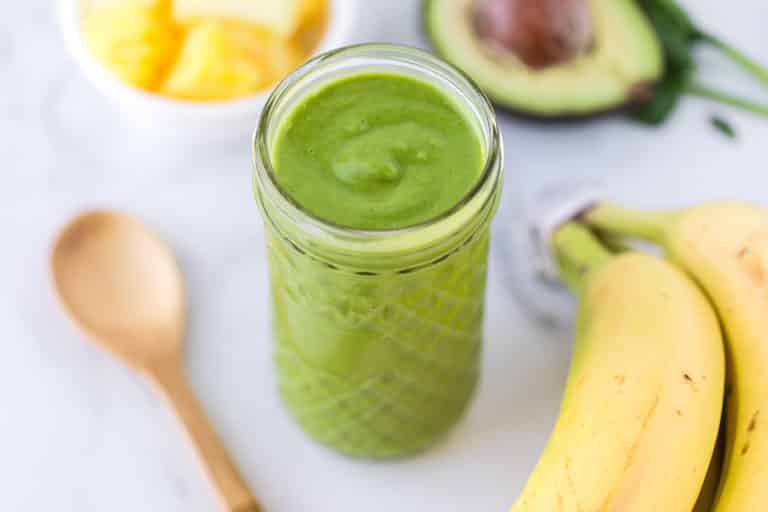 detox smoothie in a glass surrounded by a spoon, bananas, avocado, and pineapple