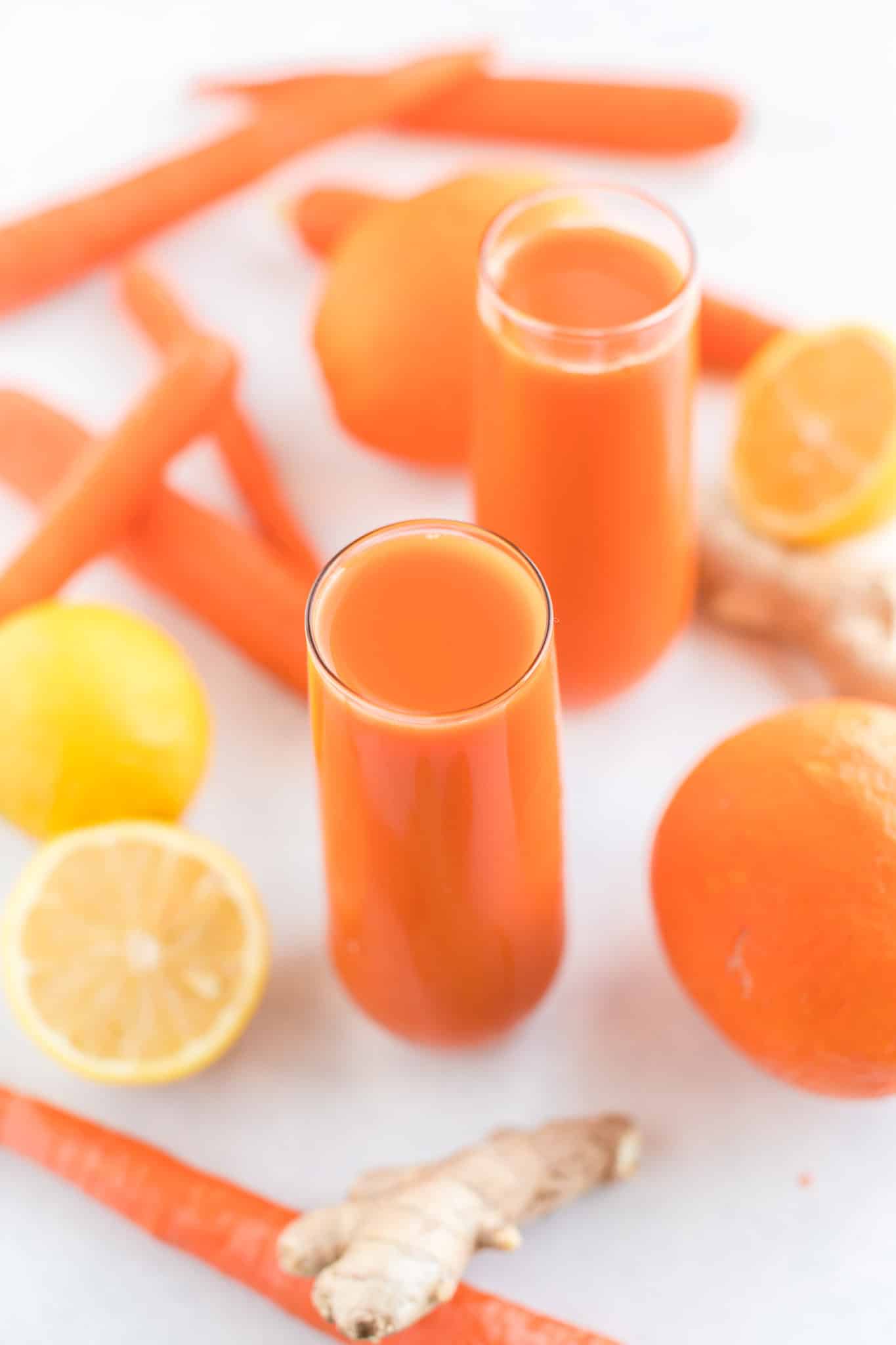 carrot juice served in two glasses on a countertop with carrots, oranges, lemons, and ginger