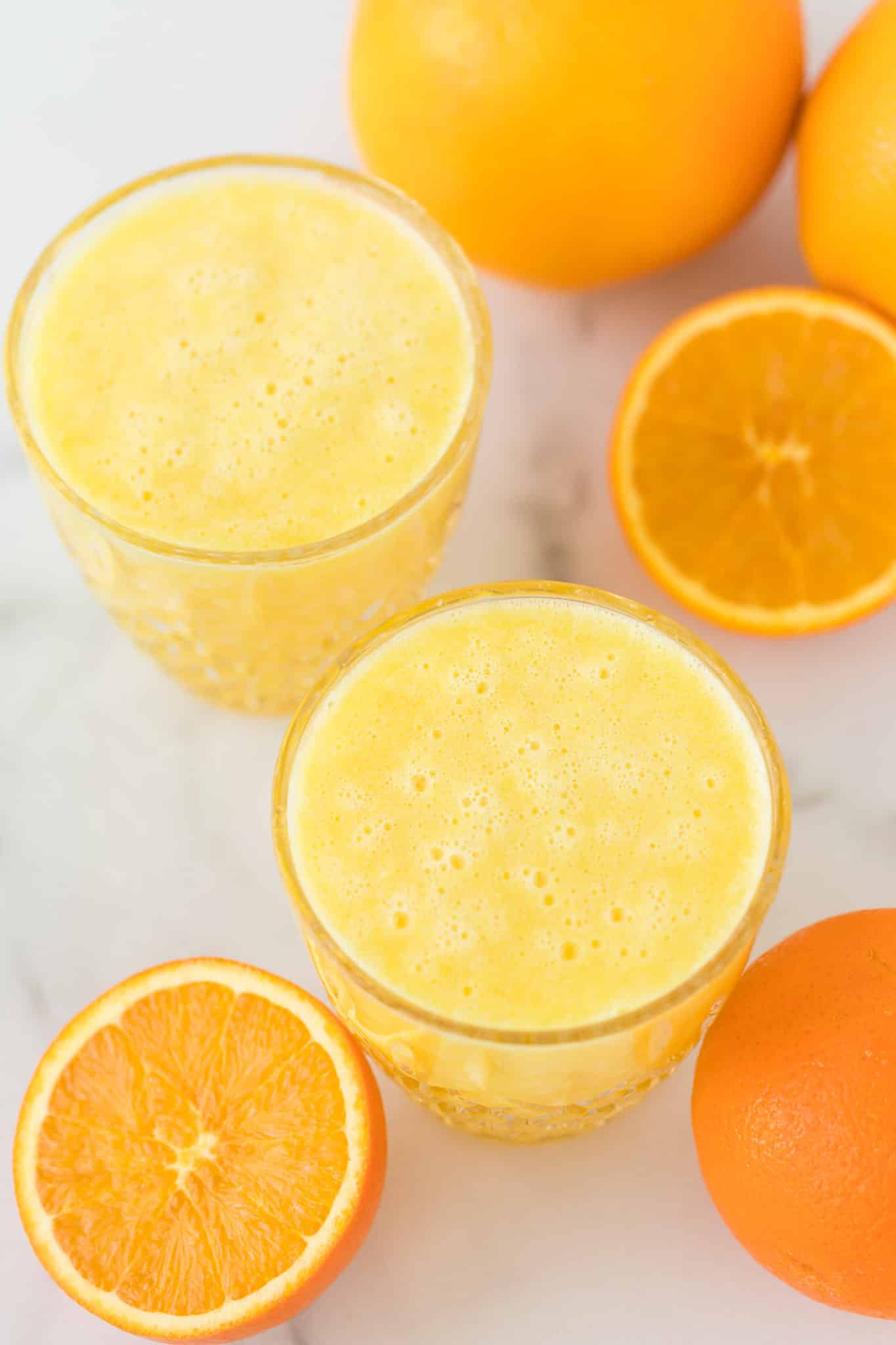 two glasses of blended orange juice on a countertop with fresh oranges