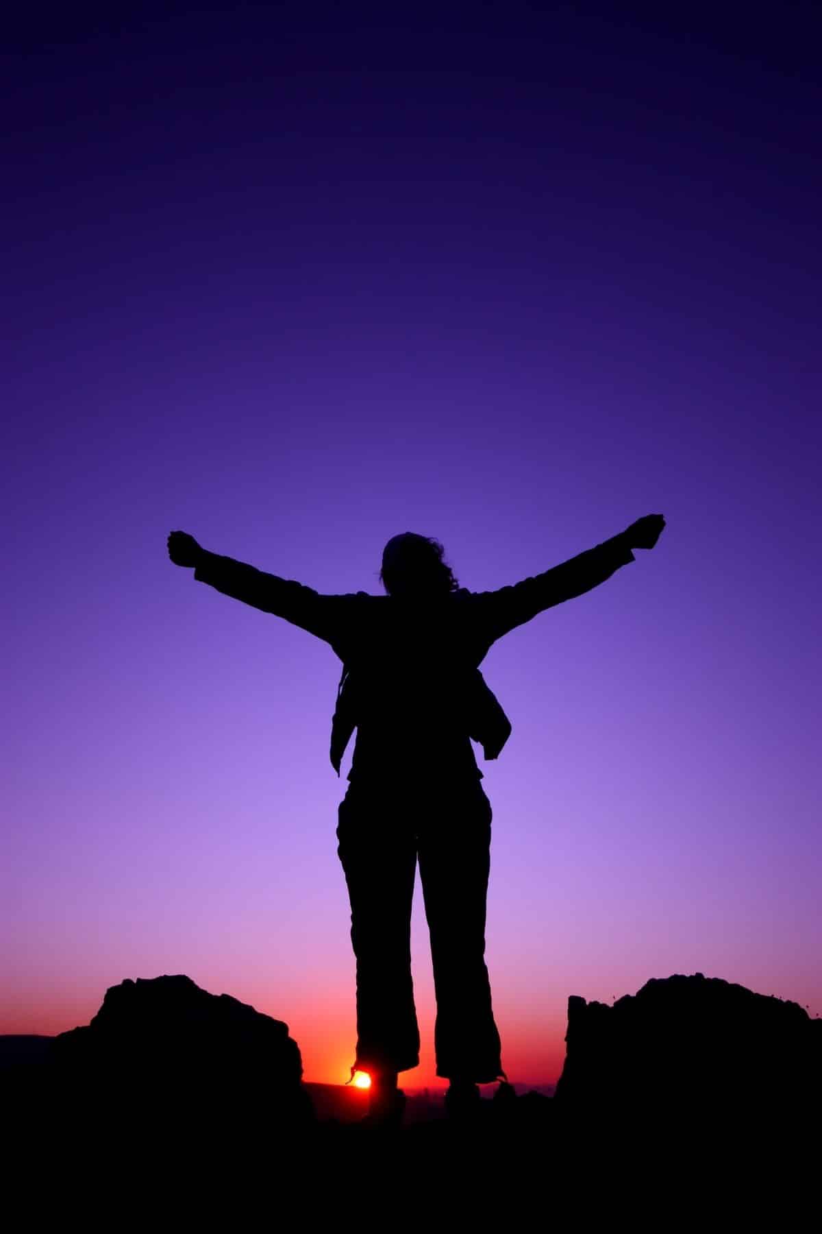 woman at sunset at the top of a mountain with her arms raised.
