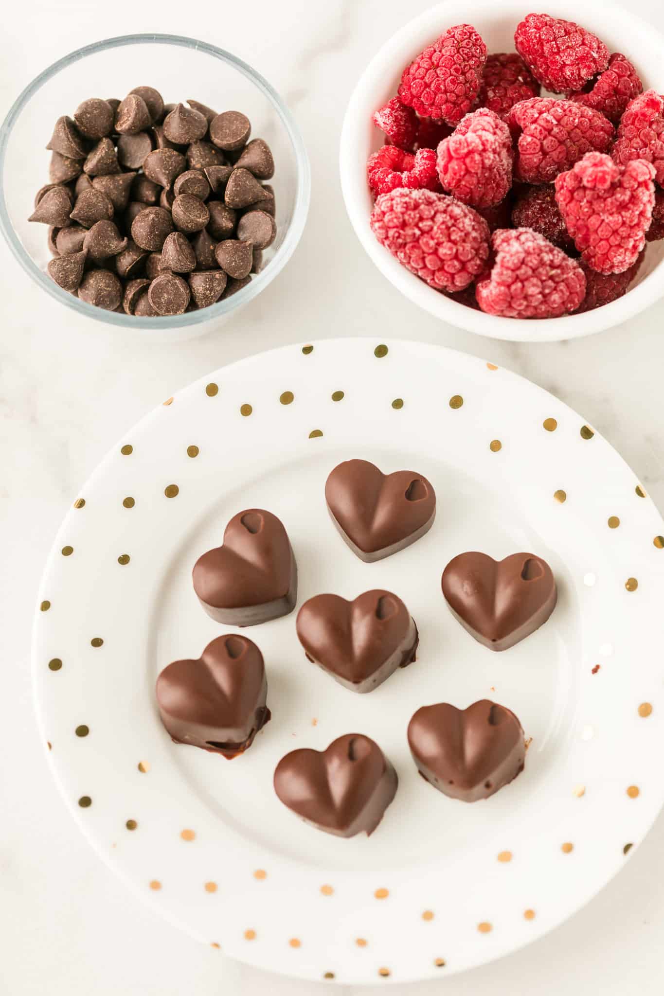 chocolate hearts on a plate next to a bowl of chocolate chips and a bowl of frozen raspberries