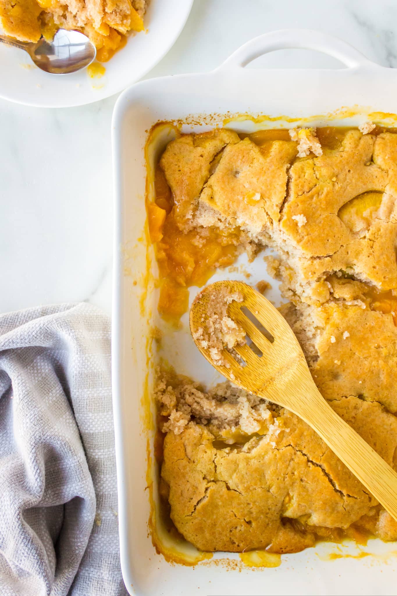 Gluten free peach cobbler in a large baking dish with a large scoop missing and a wooden spoon in it. 