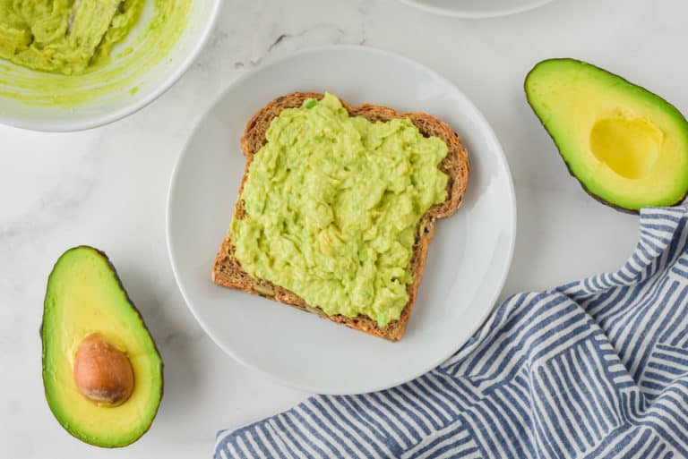 Top view of a white plate with a slice of avocado toast and a halved avocado.