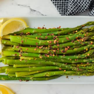 White rectangle platter of cooked asparagus with lemon wedges.