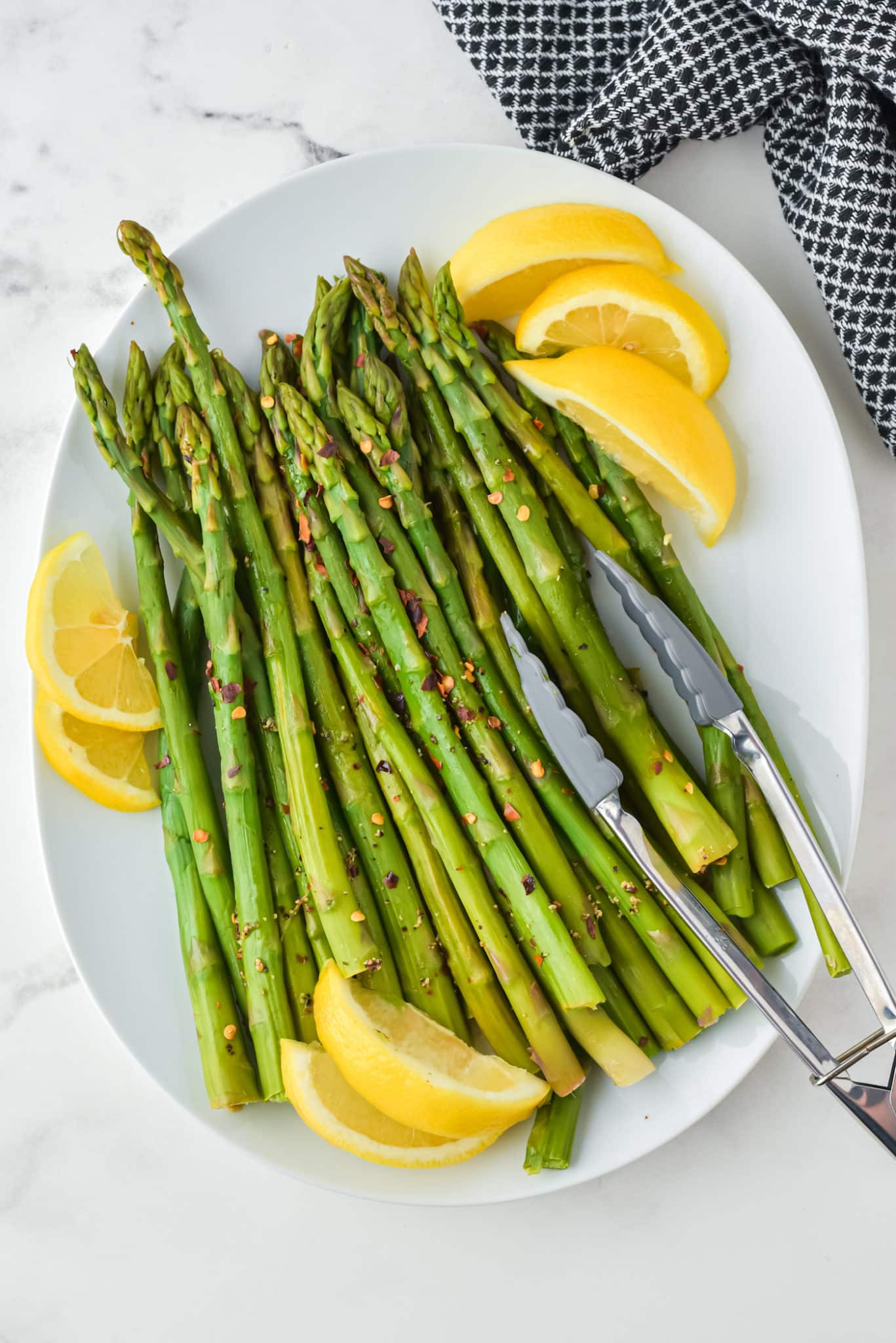 An oval white platter with cooked instant pot asparagus, lemon wedges, and tongs.