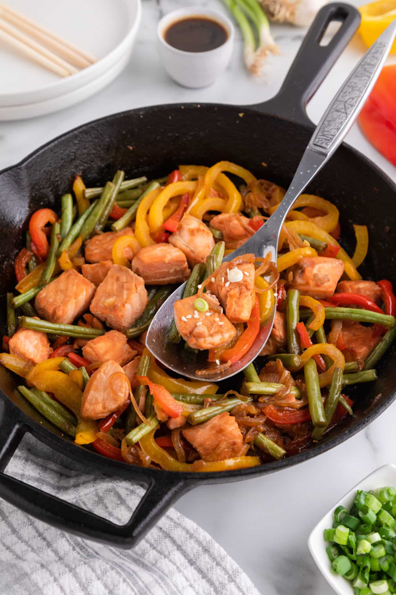 A silver serving spoon scooping salmon stir fry from a cast iron skillet.