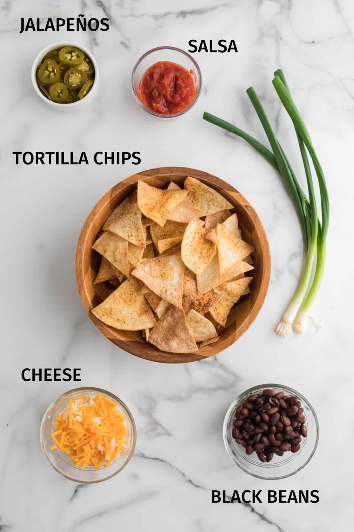 Tortilla chips and toppings for nachos in small bowls on a white surface.