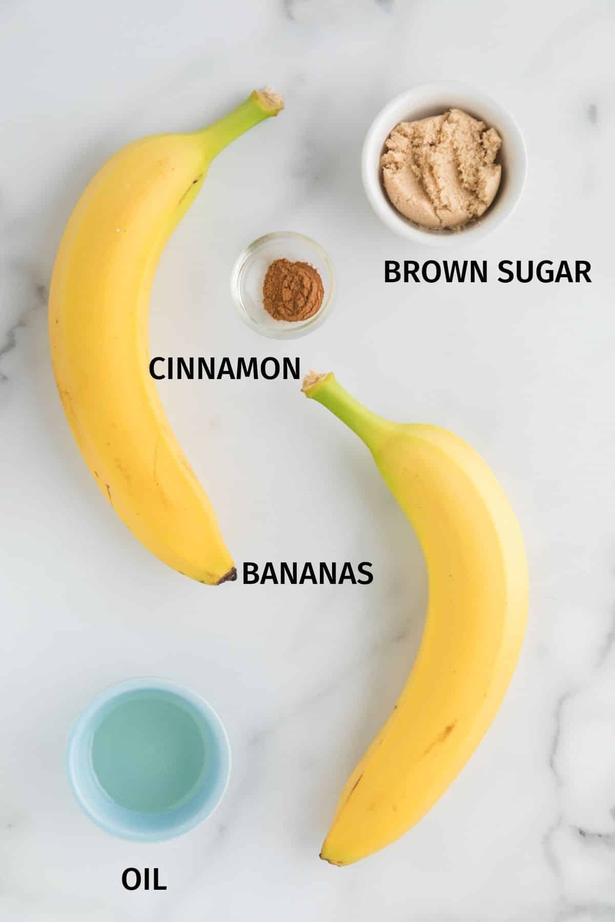 Bananas, brown sugar, cinnamon, and oil in bowls on a white surface.