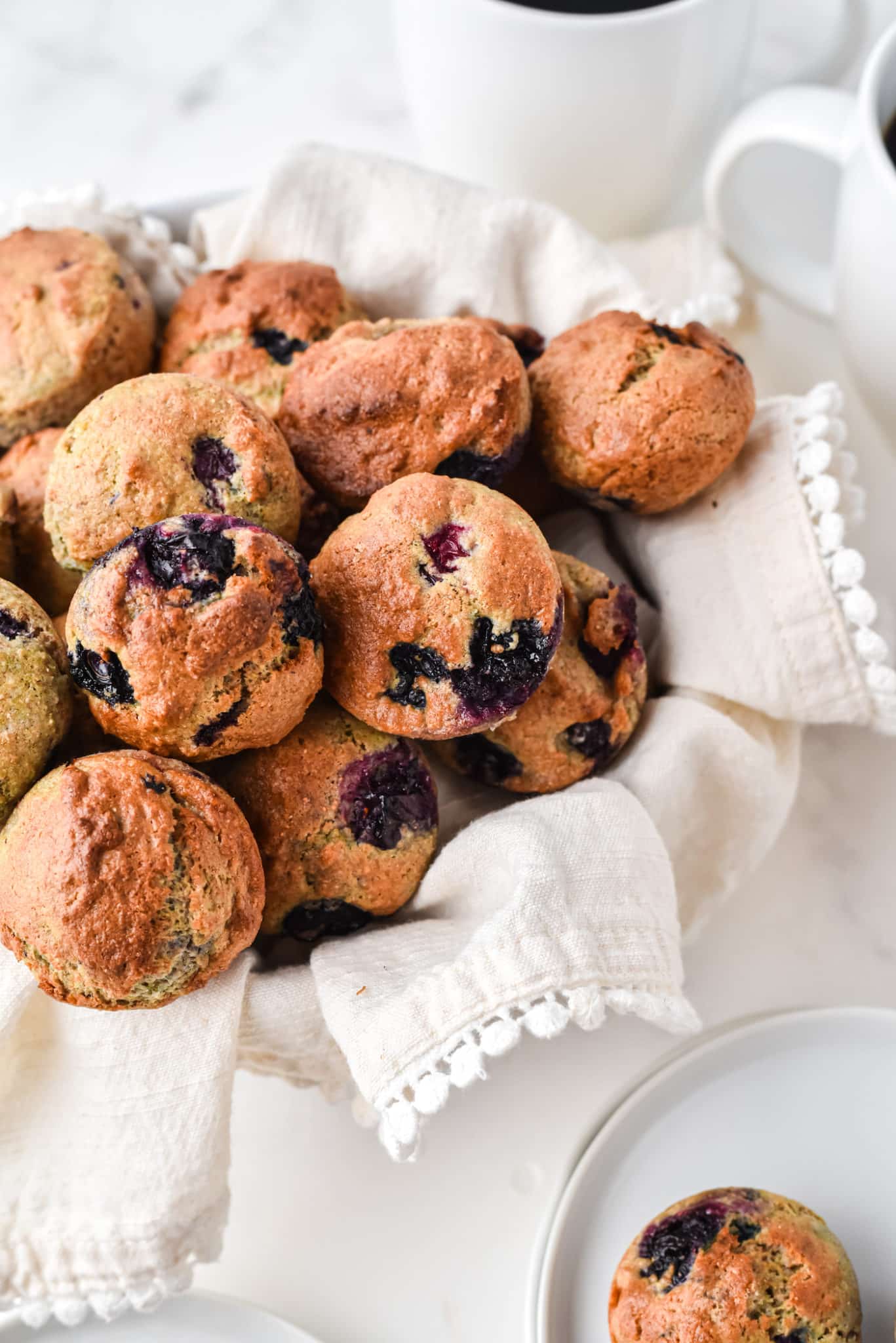 Blueberry mini muffins in a towel-lined basket next to a mug of coffee.