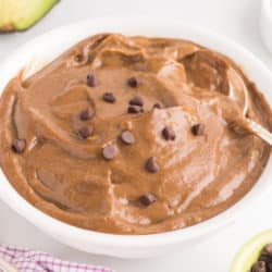 A white bowl with a spoon is filled with chocolate avocado pudding topped with chocolate chips.