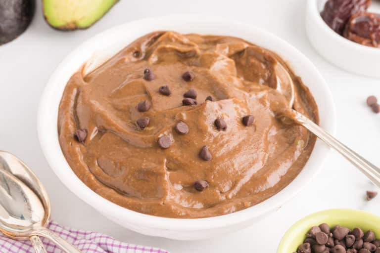 A white bowl with a spoon is filled with chocolate avocado pudding topped with chocolate chips.
