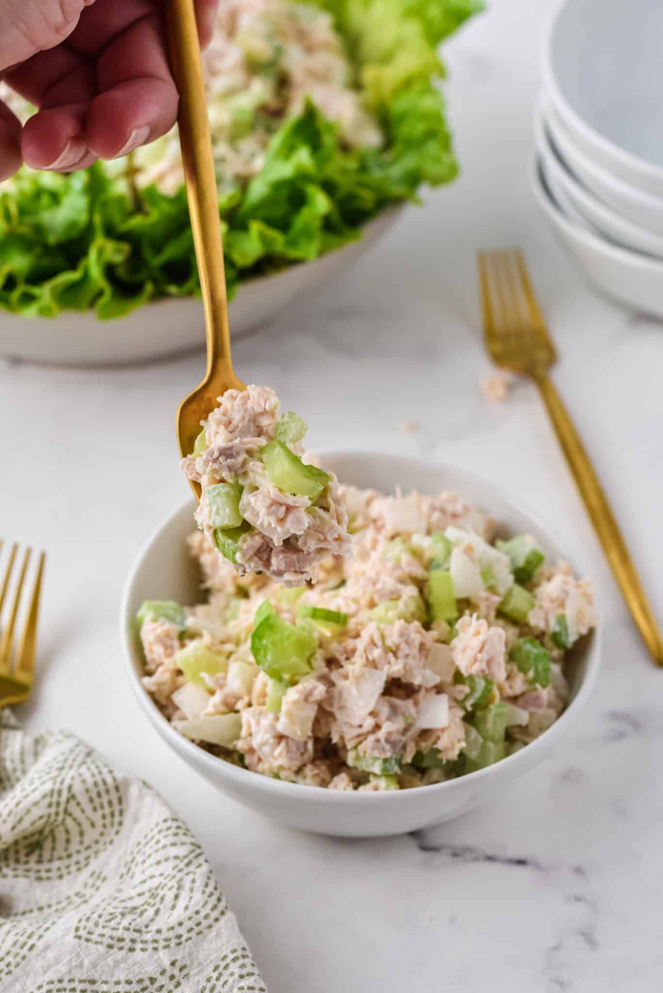 A gold fork picking up a bite of chicken salad from a small white bowl.