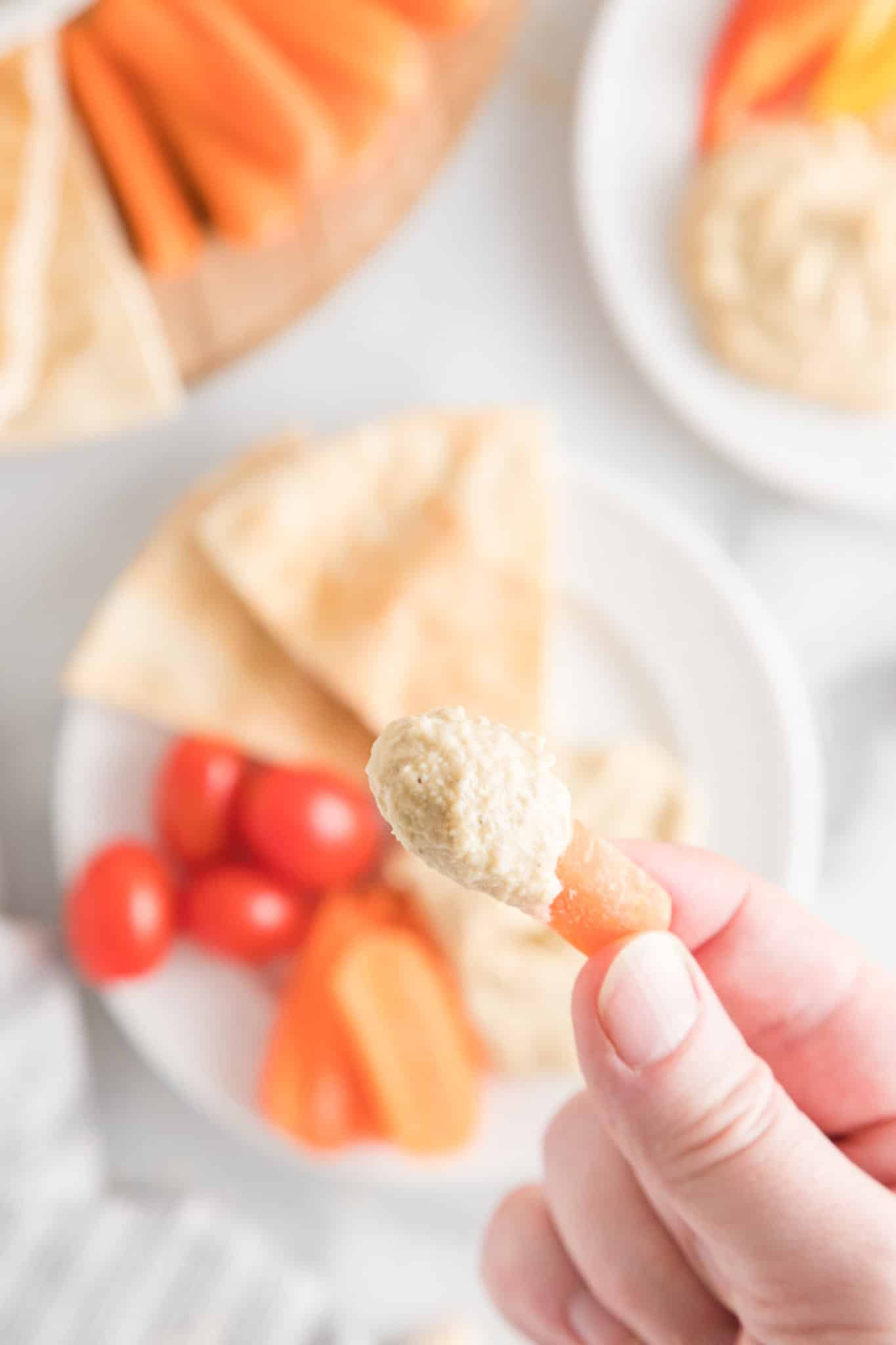 Close up of a hand holding a baby carrot that's been dipped in homemade hummus.