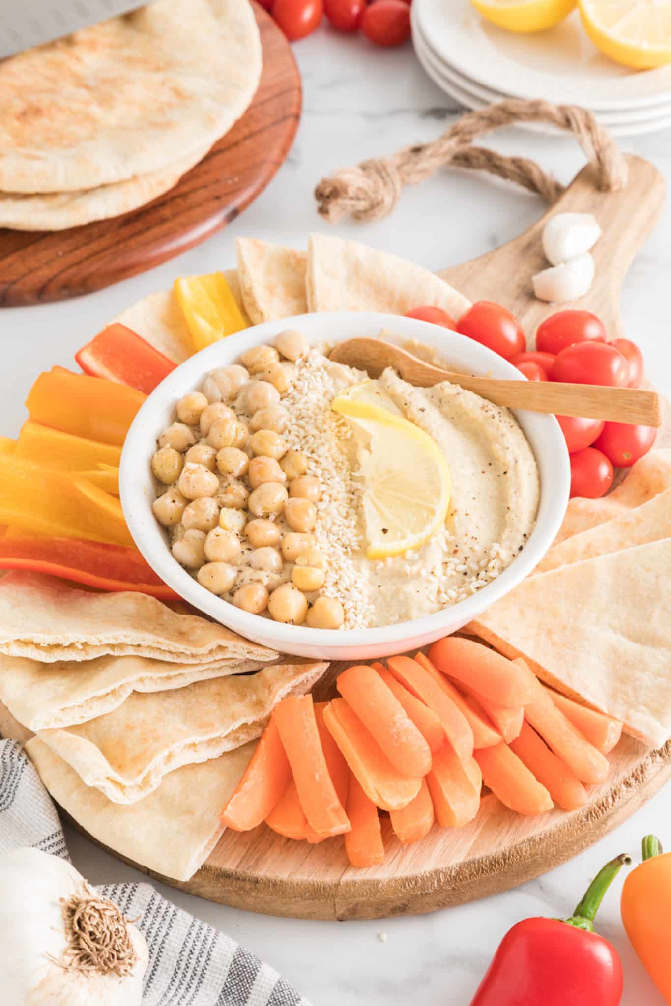 A wooden platter with sliced veggies and quartered pita around a small bowl of hummus.