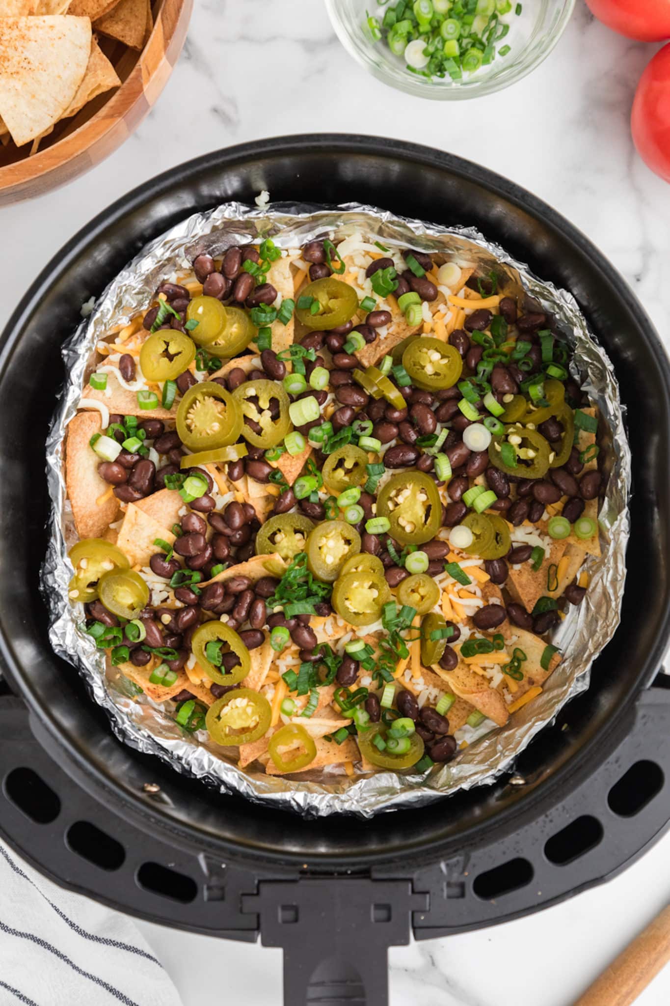 Nacho toppings of black beans and jalapenos on top of chips in an air fryer basket.