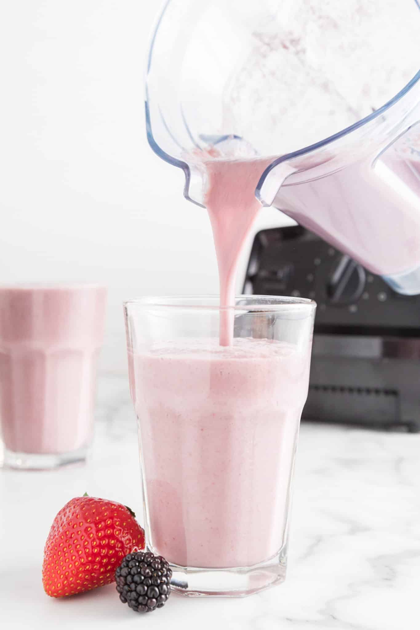 A blender jar pouring strawberry blackberry smoothie into a tall glass.