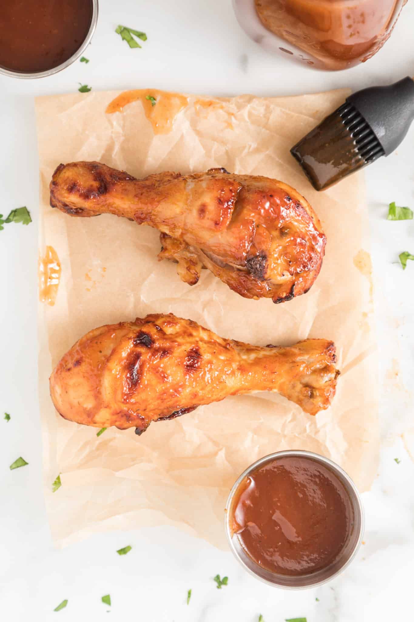 Two BBQ chicken drumsticks on parchment paper on a white surface.