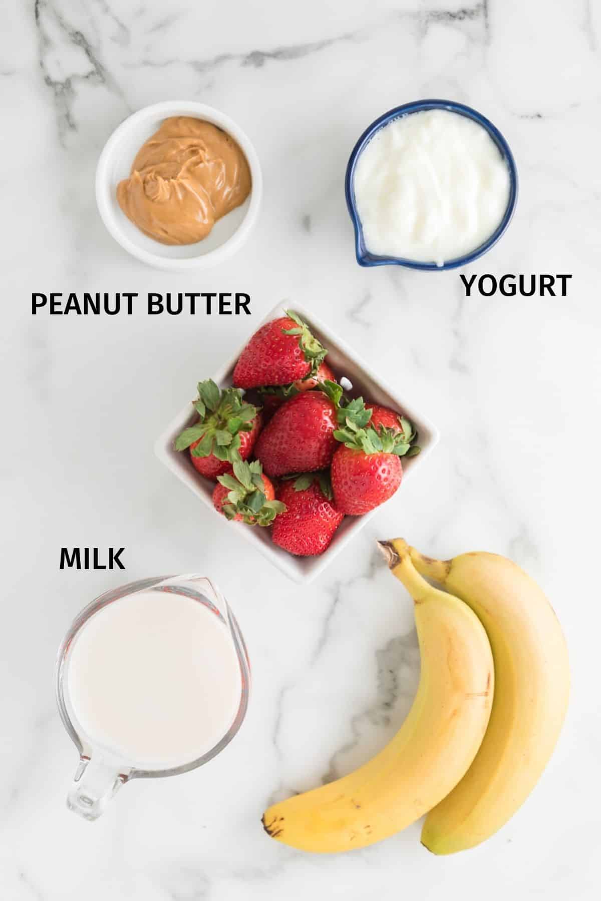 Ingredients for a strawberry banana peanut butter smoothie in small bowls on a white surface.