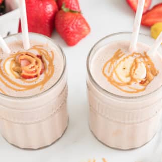 Two glasses of strawberry banana smoothie two straws and peanut butter drizzled on top.