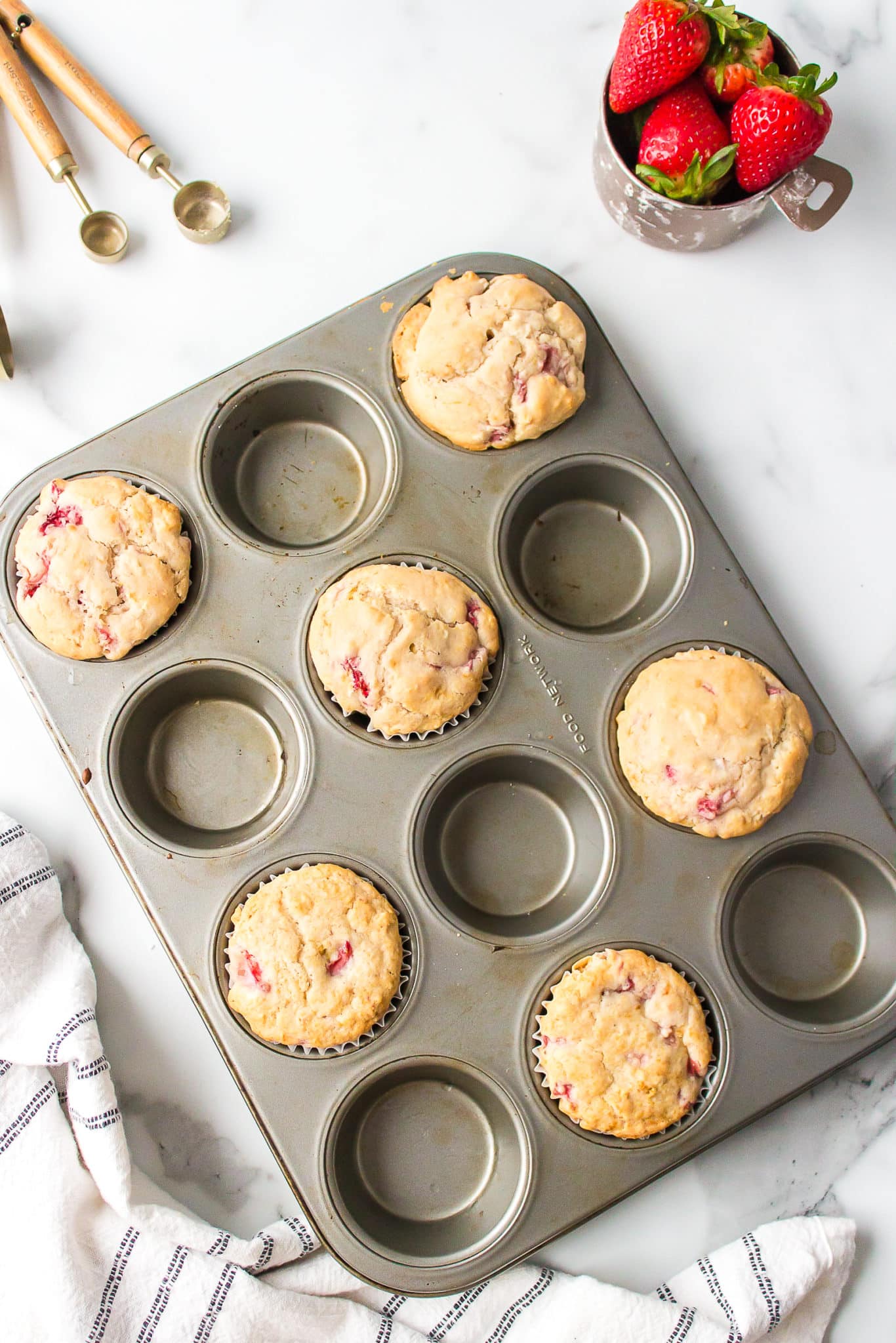Strawberry muffins in papers in a muffin tin on a white surface.