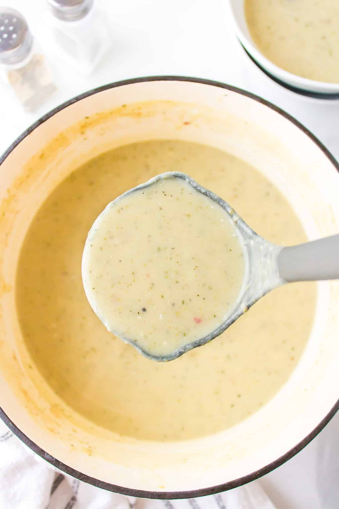 A gray ladle filled with zucchini potato soup over a Dutch oven.