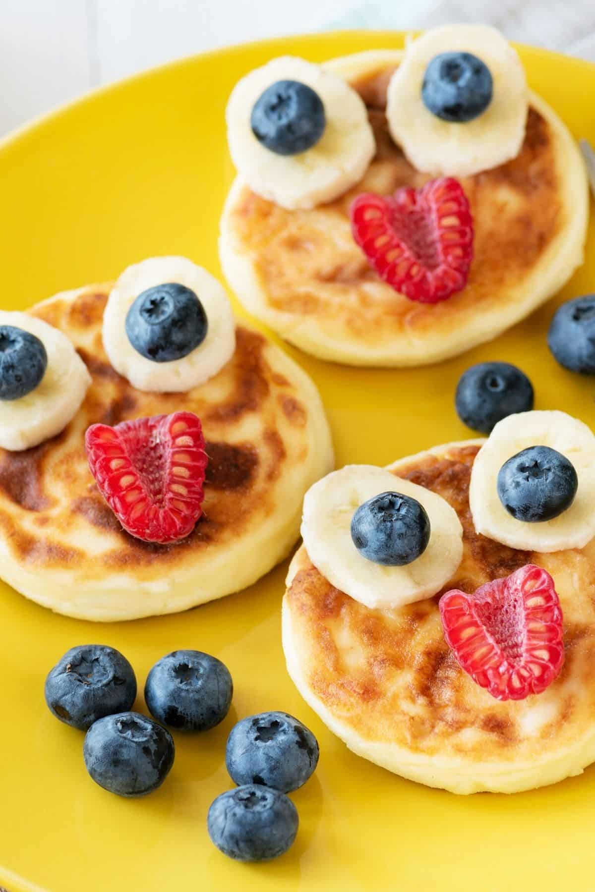 Pancakes with fruit faces on a yellow plate.