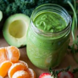 A green smoothie in a mason jar with oranges and avocado next to it.