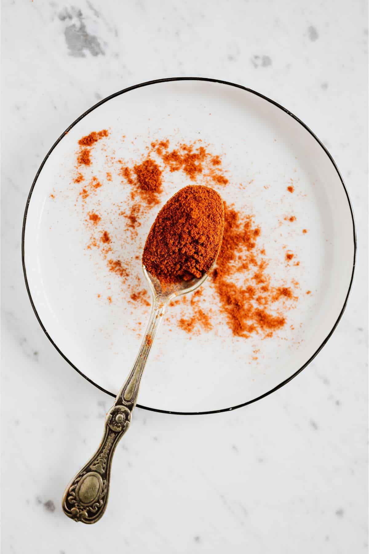 Silver spoon spreading paprika spice on white plate.