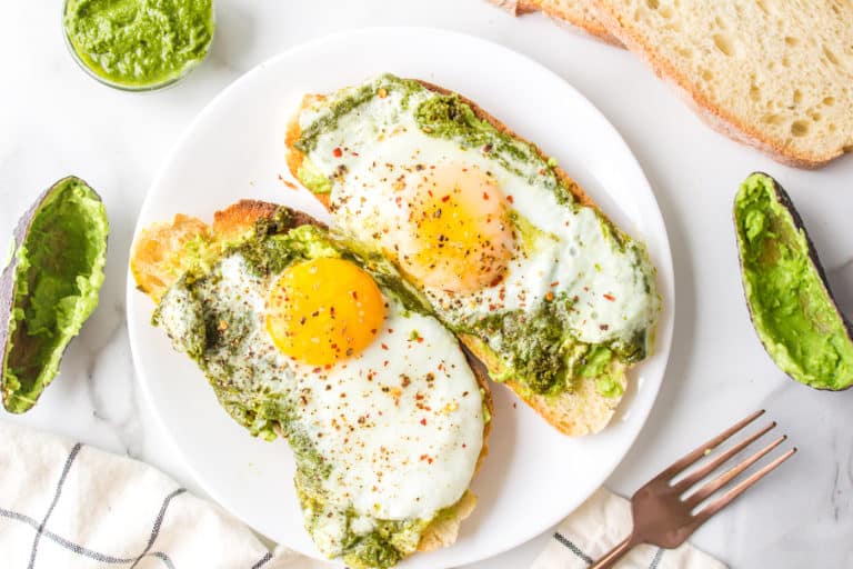 Pesto fried eggs on slices of avocado toast on a white plate.