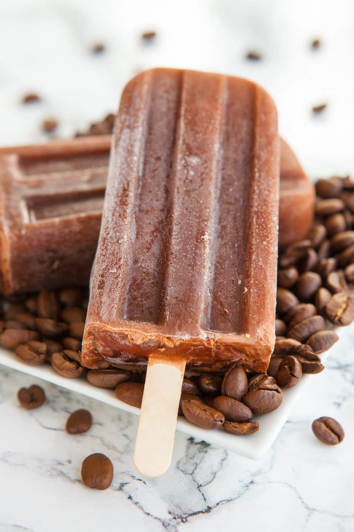 A white square plate filled with coffee beans with two coffee popsicles on top.