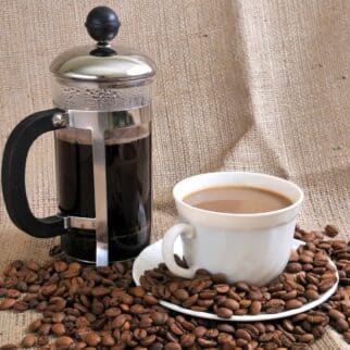 a French press and cup of coffee surrounded by coffee beans.