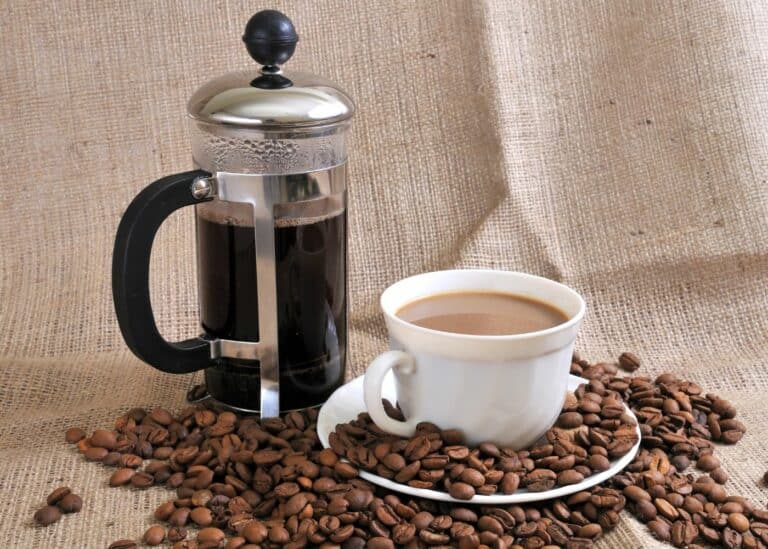 a French press and cup of coffee surrounded by coffee beans.