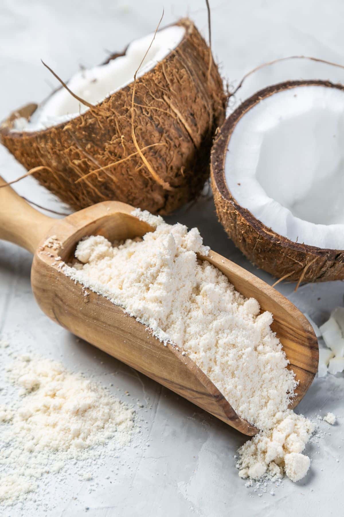 Scoop of coconut flour on wooden surface with halved coconuts.