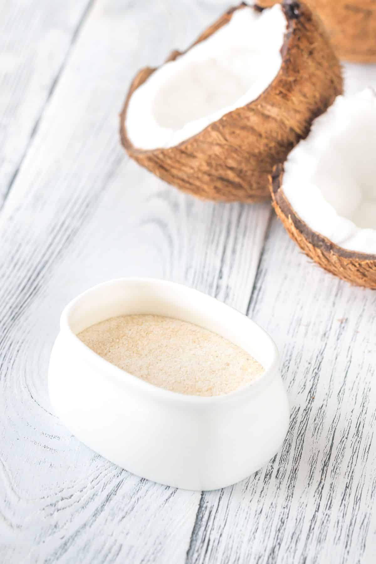 White bowl of coconut flour and coconuts on wooden surface.