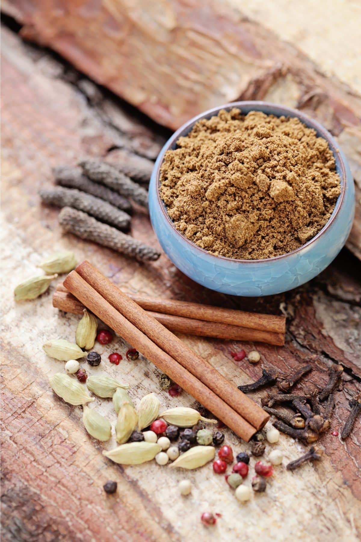 Bowl of garam masala with dried spices on wooden surface.