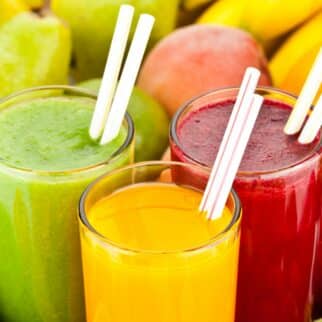 three cups of juice with straws surrounded by fruits.
