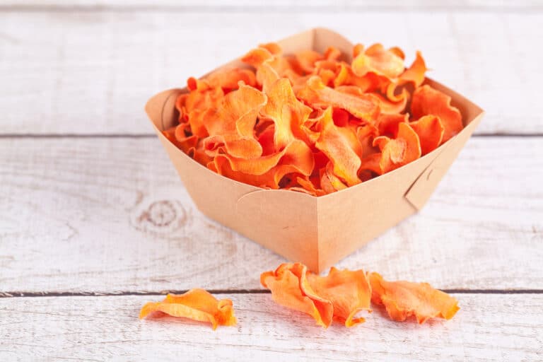 Dehydrated carrot chips in a brown box on a wooden surface.