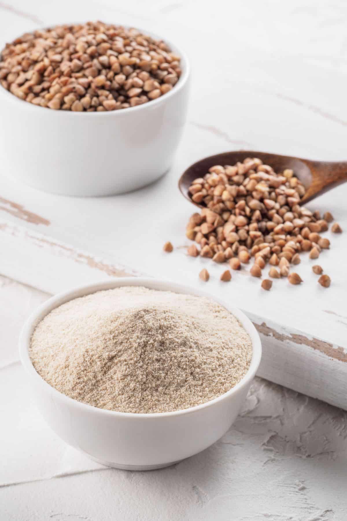 Bowls of buckwheat flour and whole buckwheat grains on white surface.