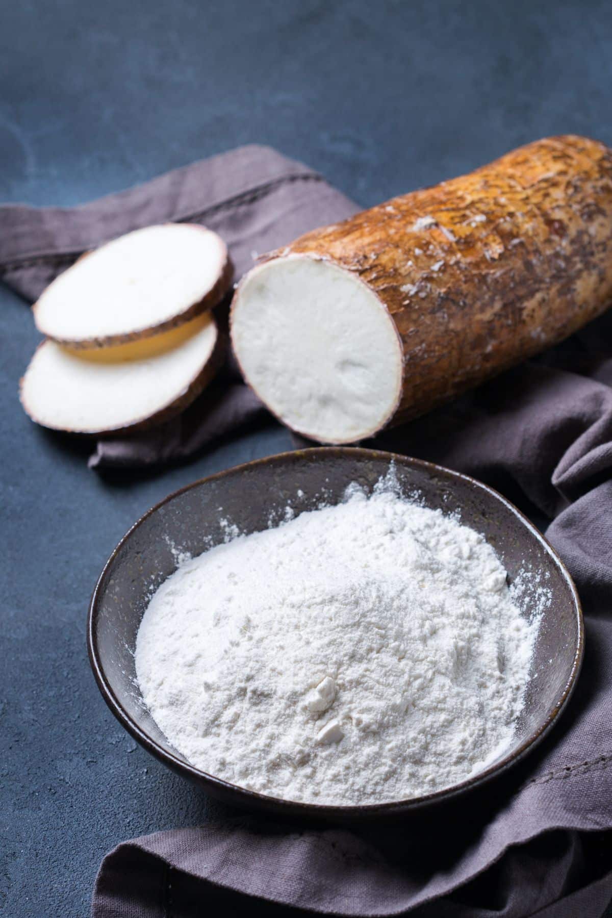 Bowl of cassava flour with cassava root on dark surface.
