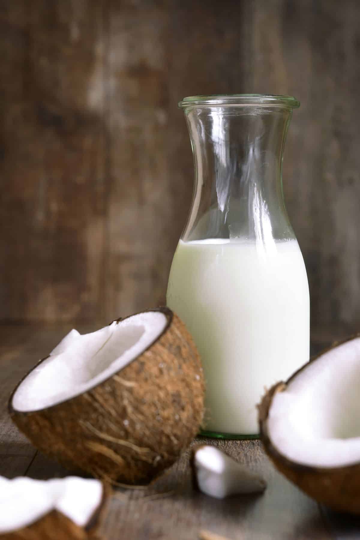 Jar of coconut milk with halved coconuts on wooden surface.