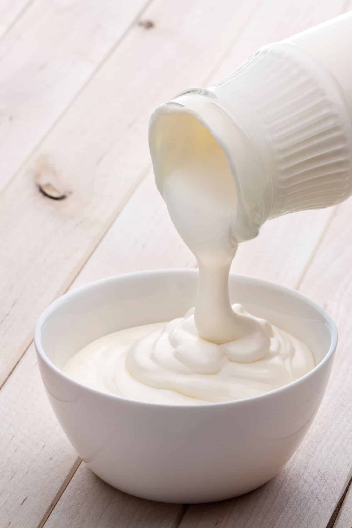 Jar pouring heavy whipping cream in white bowl on wooden surface.