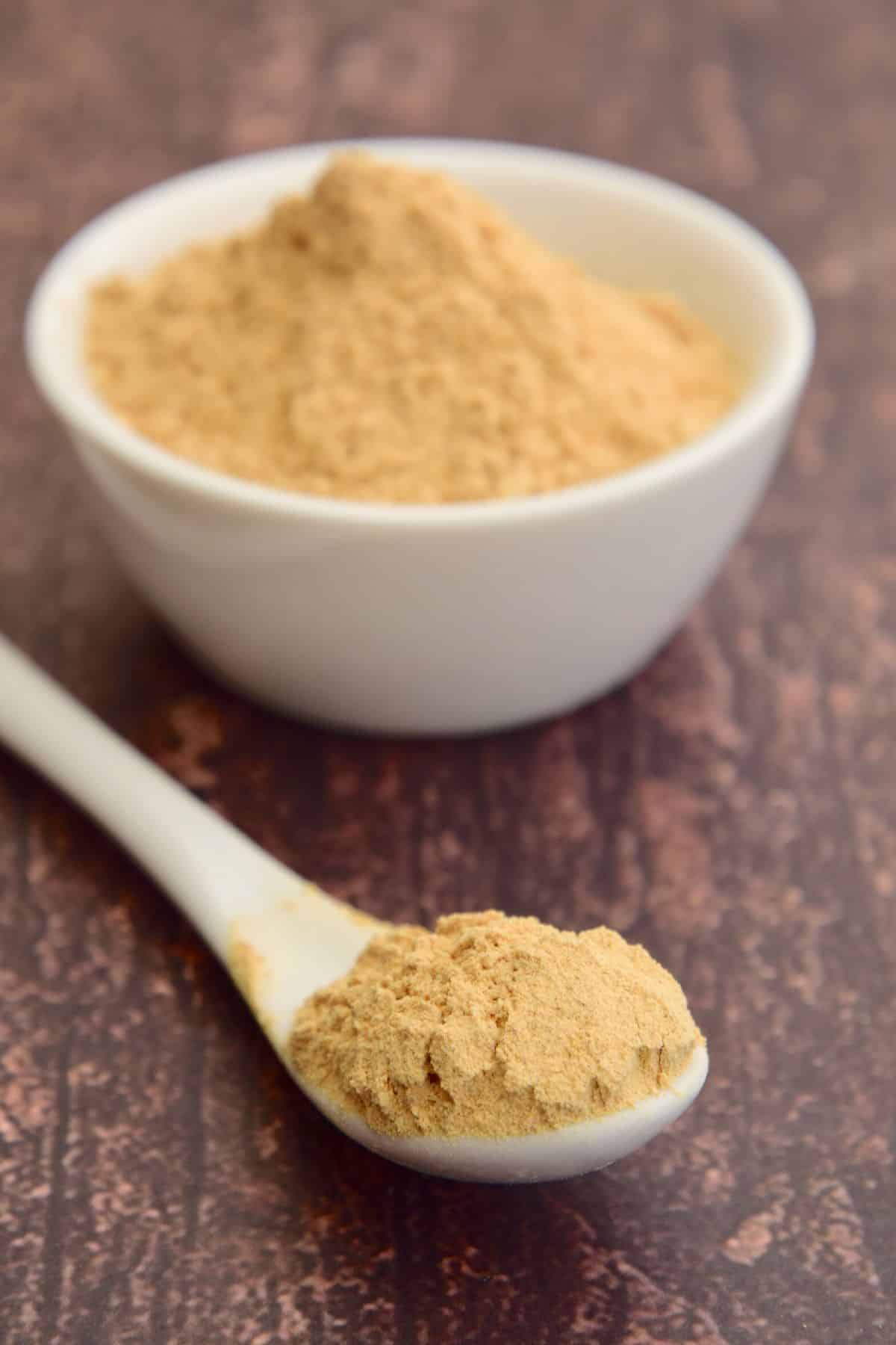 A small white spoon of maca powder next to a small bowl full.