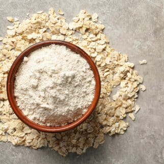 Bowl of oat flour with scattered oats on stone surface.
