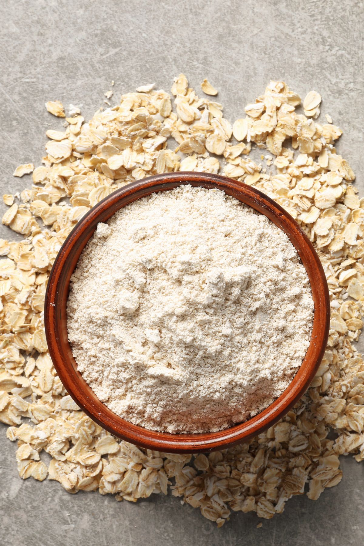Bowl of oat flour with scattered oats on stone surface.