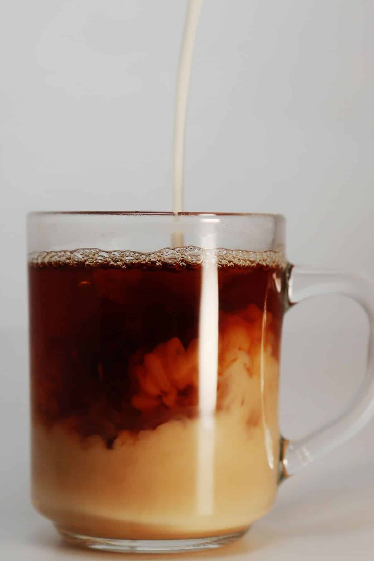 Milk being poured in a stream into a clear mug of protein coffee.