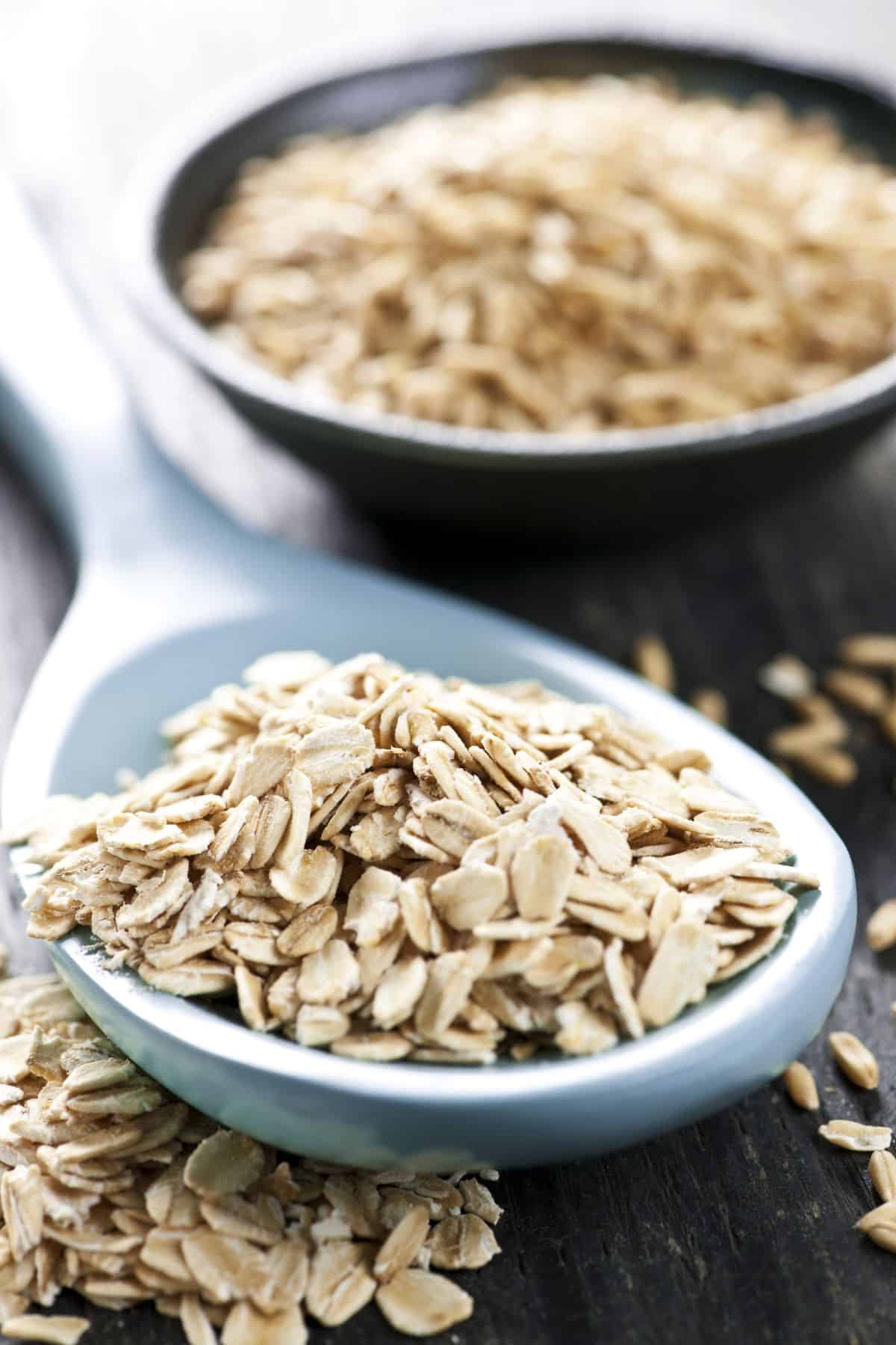 Spoonful of rolled oats with scattered oats on dark surface.